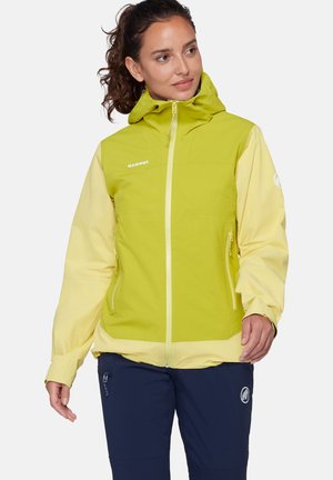 Femme portant une veste à capuche verte et jaune pâle avec des poches zippées, associée à un pantalon bleu marine avec une poche zippée et un logo.
