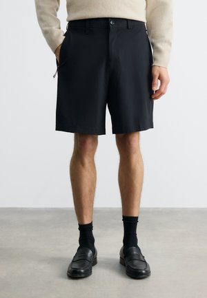 Muž oblečený v čiernych krátkych nohaviciach po kolená, čiernych ponožkách a čiernych mokasínach, stojaci na šedom podlahe s jednou rukou v vrecku.