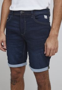 Shorts en denim sombre avec des ourlets retroussés, une coupe slim et des coutures jaunes contrastées. Comprend des poches avant et une étiquette sur la taille.