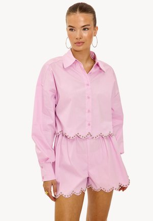 Femme portant une chemise boutonnée et un short assortis de couleur rose pâle, avec des bords festonnés décorés de petits clous métalliques.