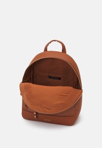 Sac à dos en cuir marron avec fermeture éclair dorée ouverte, montrant une poche intérieure et une doublure en tissu, avec une poignée de transport supérieure et une étiquette de marque « Anna Field ».