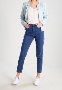 Chaqueta bomber azul claro, camiseta crema, vaqueros de tiro alto azules y zapatillas blancas. Conjunto casual con un ajuste relajado y detalles de costura visibles.