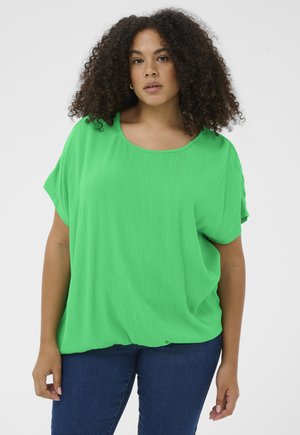 Frau mit lockigem Haar, die ein leuchtend grünes kurzärmeliges Oberteil und blaue Jeans trägt, steht vor einem schlichten weißen Hintergrund.