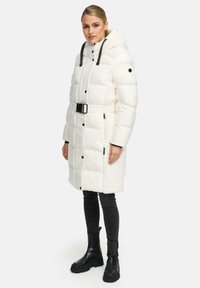 Witte pufferjas met een ceintuur in de taille, voorzien van contrasterende zwarte banden, zijzakken en een gestructureerd paneel. Model draagt zwarte jeans en laarzen.