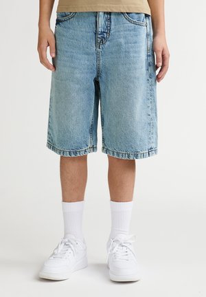 Pantaloni scurți din denim albastru deschis, cu croială lejeră, lungime până la genunchi, având tiv răsfrânt și design clasic cu cinci buzunare. Purtați cu adidași albi și șosete.