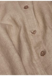 Leinenhemd in hellem Beige mit sichtbarer Struktur. Verfügt über braune Knöpfe, einen strukturierten Kragen und eine Knopfleiste mit sichtbaren Nähdetails.