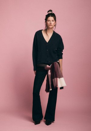 Schwarze Strickjacke mit großen Knöpfen, kombiniert mit dunklen Schlagjeans. Ein mehrfarbiges Tuch ist um die Taille gewickelt. Pinker Hintergrund.