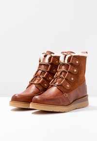 Un par de botas de invierno de cuero y ante marrón con forro de piel, cintas de encaje y suelas de goma sobre un fondo blanco.