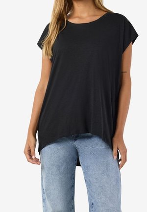 Femme portant un haut noir à manches courtes et amples, et un jean taille haute bleu clair, debout devant un fond blanc.