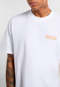 Bílé bavlněné tričko s krátkými rukávy, kulatým výstřihem a oranžovým logem potištěným na levé straně hrudi. Hladký materiál, uvolněný střih.