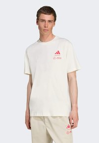 Bílá bavlněná tričko s kulatým výstřihem, na levé straně hrudi s růžovými logy Adidas a Mercedes. Krátké rukávy a volný střih.