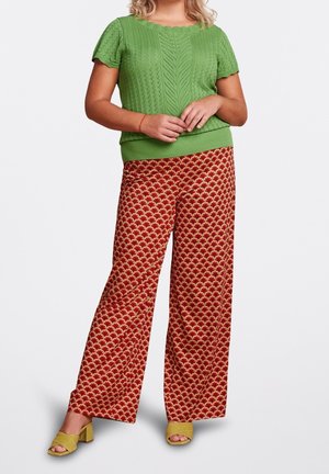 Femme portant un top en maille vert à manches courtes, un pantalon rouge large à motifs, et des sandales jaunes à talons carrés et bout ouvert, debout, les mains jointes.