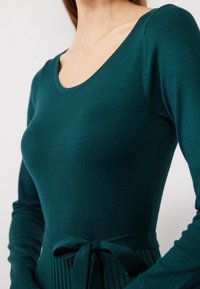 Abito teal a maniche lunghe realizzato in tessuto a coste, con corpetto aderente e cintura in vita. La gonna presenta pieghe verticali per texture.