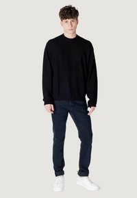 Maglione nero a collo rotondo con design texturizzato, abbinato a jeans blu scuro e sneakers bianche, in piedi su uno sfondo grigio chiaro.