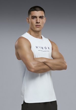 Hombre musculoso con cabello corto cruzando los brazos, vistiendo una camiseta blanca sin mangas con el texto "HYROX", de pie frente a un fondo gris liso.