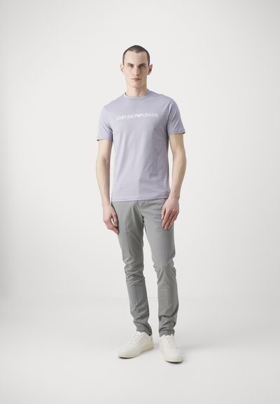 Heleda lilla T-särk, millel on "EMPORIO ARMANI" logo, kokku sobitatud hallide slim-fit pükste ja valgete spordijalatsitega. Sujuv kangas, kaasaegne lõige, minimaalne disain.