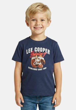 Jeune garçon souriant aux cheveux blonds portant un t-shirt bleu marine avec un motif de gants de boxe et du texte, associé à un jean bleu.