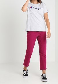 Grijze T-shirt met korte mouwen en een blauw-rood "Champion" logo, gecombineerd met een roze straight-leg broek en zwarte sneakers.