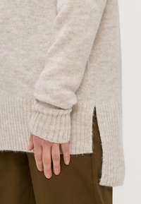 Beige strikkegenser med ribbestrikkede mansjetter og sidespalter, laget av mykt, teksturert stoff. Hånden hviler på olivengrønne bukser.