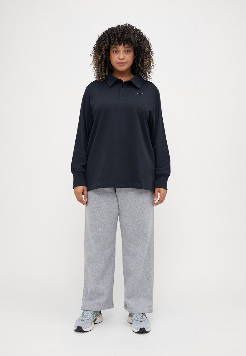 Polo noir à manches longues avec col et logo brodé, associé à un pantalon de survêtement gris à jambe large et des chaussures de sport multicolores.