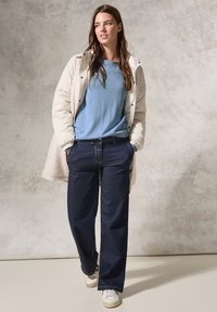 Beige puffjacka lager över en ljusblå skjorta, parat med mörkblå vida jeans och vita sneakers, mot en texturerad bakgrund.