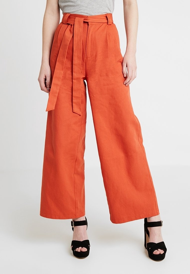 Pantalon orange taille haute à jambes larges pour femme, avec une ceinture assortie en tissu, associé à des sandales à talons hauts noires ouvertes et un haut gris.