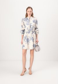 Rochie scurtă cu imprimeu floral în crem și albastru, având mâneci bufante, un decolteu cu butoni și o talie ajustabilă cu un cordon legat. Poartă o geantă de mână argintie și tocuri.