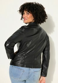 Veste en cuir noir au design ajusté, avec des poignets zippés et des coutures courbes. Associée à un jean en denim bleu clair et présentée sur un fond neutre.