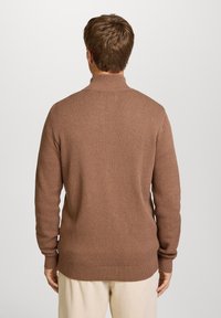Brun strikket sweater med høj krave, struktureret mønster, lange ærmer samt ribbede manchetter og kant, båret med lyse bukser.