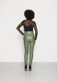 Mujer con cabello rizado vistiendo una blusa negra transparente de manga larga, pantalones de cuero verde oliva de cintura alta y sandalias negras de tacón alto, de espaldas.