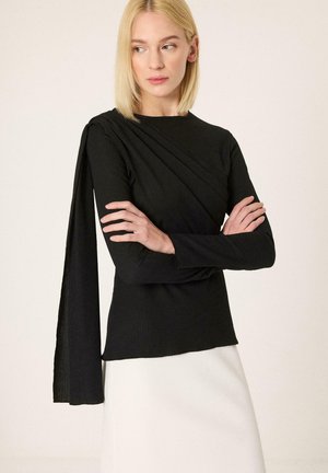 Ξανθιά γυναίκα με ίσια μαλλιά φορά μαύρο μακρυμάνικο τοπ με λεπτομέρεια draped υφάσματος στον ένα ώμο και λευκή φούστα, χέρια σταυρωμένα.