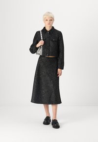 Crni tweed sako s rezom do struka i odgovarajuća duga suknja, koja ima suptilan metalik sjaj. Uparen s crnim cipelama na vezanje.