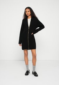 Μαύρη ριμπ πλεκτή ζακέτα με φερμουάρ στο μπροστινό μέρος, μακριά μανίκια και τσέπες. Διαθέτει χρυσό φερμουάρ, σχέδιο μέχρι το γόνατο και απαλό πλεκτό ύφασμα.