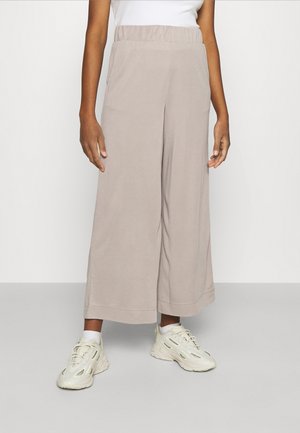 Osoba ubrana w beżowe, szerokie spodnie o skróconej długości, białe skarpetki, kremowe masywne sneakersy oraz biały top bez rękawów, stojąca na tle jednolitej ściany.