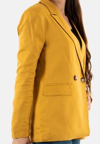 Marynarka w kolorze musztardowej żółci o dopasowanym kroju, z dwoma guzikami, klapami z wycięciem i pojedynczą kieszenią na piersi. Wykonana z tkaniny o fakturze.