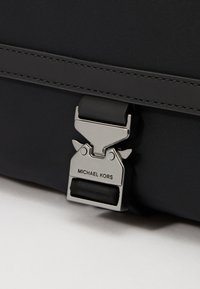 Borsa nera in nylon con un ganci in metallo argento con "MICHAEL KORS" inciso, texture liscia e dettagli cuciti a contrasto.
