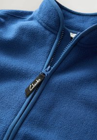 Giacca con zip a un quarto blu in pile, caratterizzata da una superficie testurizzata, zip liscia con un'etichetta "Clarks" in bianco e nero e un design con colletto alto.