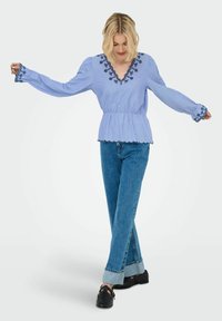 Blouse peplum bleu clair avec un col en V, motif floral noir brodé et manches évasées. Associée à un jean bleu taille haute et des chaussures noires.