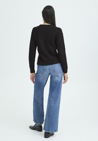 Kvinde med langt mørkt hår står med ryggen til iført en tekstureret sort sweater, blå vide jeans, grå sokker og sorte sko.