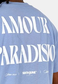Blauwe katoenen t-shirt met grote witte tekst: "AMOUR PARADISIO" op de achterkant, met een klein logo en tekst onderaan. Eenvoudig ontwerp met ronde hals.