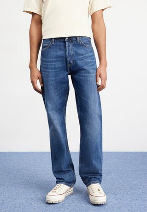 Homme portant un jean bleu coupe décontractée, des baskets blanches et une chemise beige clair à manches courtes, debout sur un tapis bleu.