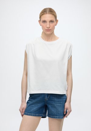 Mujer con cabello rubio recogido en una coleta baja, lleva una camiseta blanca sin mangas y shorts vaqueros azules, de pie frente a un fondo blanco liso.