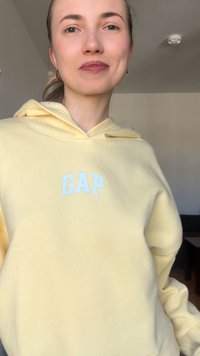Νεαρή γυναίκα με ανοιχτόχρωμο δέρμα και μαλλιά δεμένα πίσω φοράει ανοιχτό κίτρινο φούτερ GAP, χαμογελώντας ελαφρώς, σε εσωτερικό χώρο με ουδέτερο φόντο.