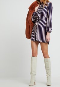 Pruhované šaty s dlouhými rukávy v námořnické modři a smetanové barvě, doplněné hnědým kabátem z umělé kožešiny. Modelka má na sobě krémové boty po kolena.