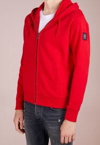 Felpa rossa con zip realizzata in tessuto morbido, dotata di tasca frontale, polsini e orlo a costine. Presenta un cappuccio con lacci e una patch con il logo sulla manica.