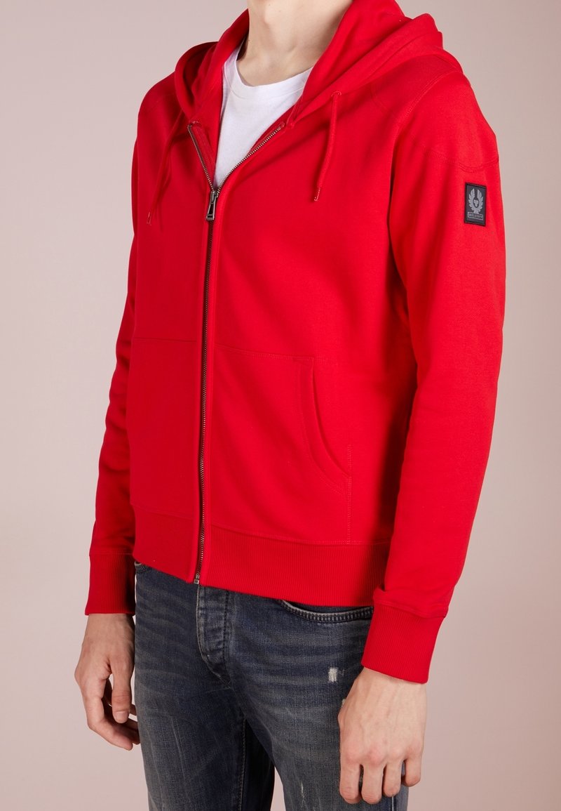 Felpa rossa con zip realizzata in tessuto morbido, dotata di tasca frontale, polsini e orlo a costine. Presenta un cappuccio con lacci e una patch con il logo sulla manica.