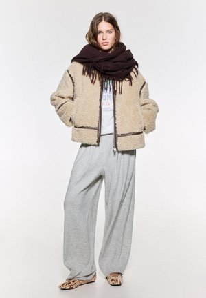 Giacca sherpa beige con rifiniture marrone scuro, pantaloni della tuta grigi oversize e scarpe slip-on con motivo; abbinata a una sciarpa marrone scuro.