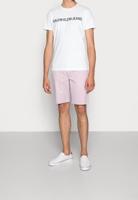 Bílý bavlněný tričko s černým potiskem "CALVIN KLEIN JEANS", světle růžové kraťasy a bílé boty na slip-on s hnědými akcenty.