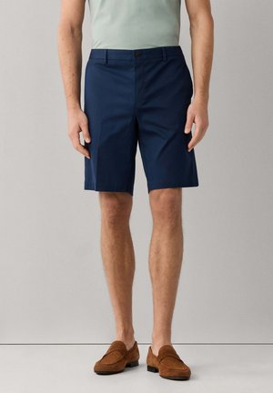 Mann trägt marineblaue maßgeschneiderte Shorts, hellgrünes Hemd und braune Wildleder-Loafer, steht vor einem schlichten hellen Hintergrund.