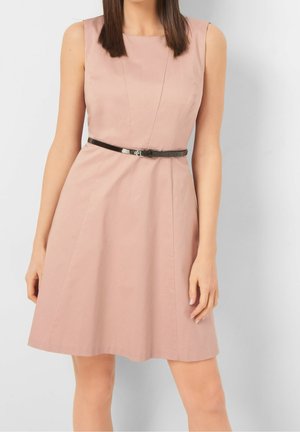 Vrouw draagt een mouwloze lichtroze jurk met een dunne zwarte riem, staande tegen een effen witte achtergrond.